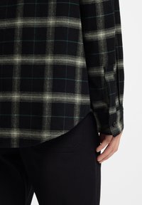 Πουκάμισο με καρό μοτίβο σε μαύρο και κρεμ με μπλε λεπτομέρειες, διαθέτει μανσέτα με κουμπιά και ελαφρώς πιο μακρύ πίσω τελείωμα, κατασκευασμένο από απαλό ύφασμα.