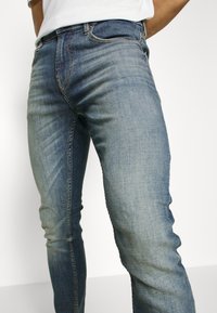 Vaqueros de mezclilla azul claro con un corte ajustado, que presentan áreas descoloridas, diseño clásico de cinco bolsillos y detalles de costura clara.