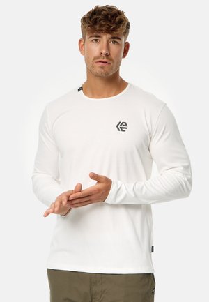 White long-sleeve shirt made of soft fabric with a round neckline and a black logo on the chest, paired with olive green pants.