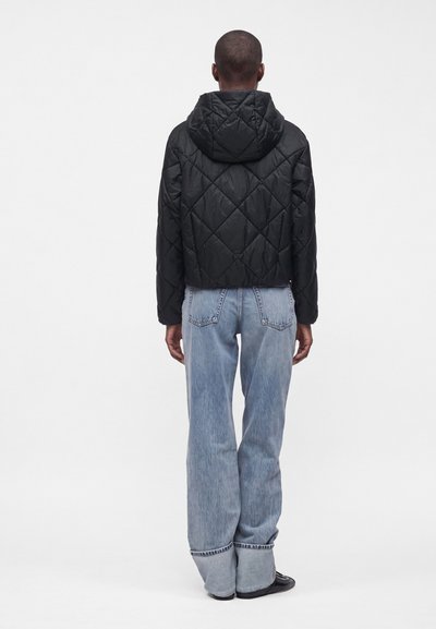 Personne debout de dos portant une veste noire matelassée à capuche et un jean bleu clair avec revers, ainsi que des chaussures noires sur un fond uni.