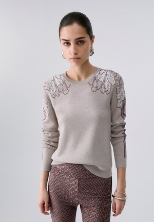 Giovane donna con i capelli pettinati all'indietro che indossa un maglione beige chiaro con ricamo floreale e pantaloni marroni a fantasia su uno sfondo neutro.