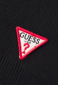 Tessuto nero a righe con una toppa triangolare ricamata in rosso e bianco con la scritta "GUESS USA" e un punto interrogativo rosso.
