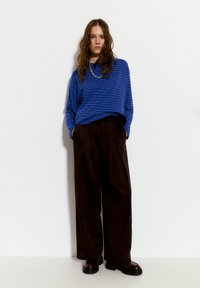 Modrý pruhovaný dlouhý rukáv spárovaný s vysokými hnědými kalhotami širokého střihu, s hladkou texturou a černými robustními botami.