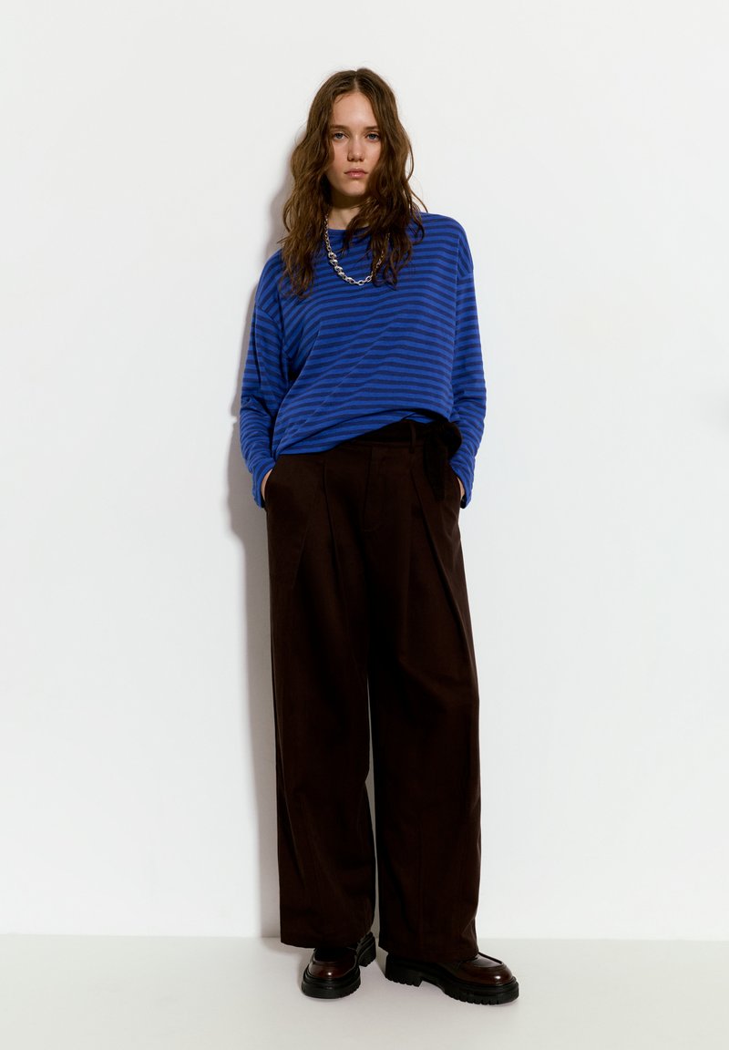 Modrý pruhovaný dlouhý rukáv spárovaný s vysokými hnědými kalhotami širokého střihu, s hladkou texturou a černými robustními botami.