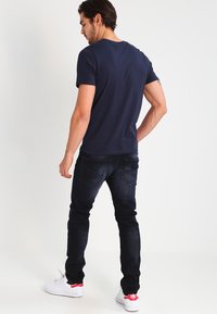 Marineblauwe t-shirt met korte mouwen gecombineerd met donkerblauwe jeans en witte sneakers. Het t-shirt heeft een ronde hals; de jeans hebben achterzakken en een slim fit.