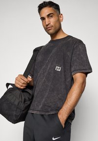 Homme portant un T-shirt noir délavé avec un petit logo, un pantalon de jogging noir Nike, tenant la bandoulière d'un sac de sport noir sur l'épaule, une main dans la poche.