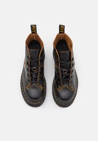 Botas de cuero negras con acentos marrones, costuras amarillas, punta redonda, diseño con cordones y una pestaña en el talón para facilitar su puesta.