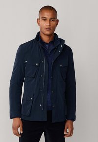 Ung mand iført en marineblå zip-up jakke med flere klaplommer over en blå trøje og skjorte, der står foran en ensfarvet baggrund.