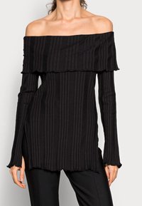 Žena má na sobě černý žebrovaný top s dlouhým rukávem a širokými manžetami, který odhaluje ramena, doplněný o černé kalhoty. Top má mírně volánkový lem.