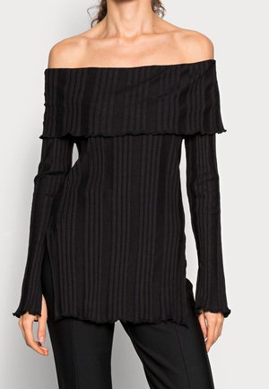 Eine Frau trägt ein schwarzes, geripptes, schulterfreies Langarmoberteil mit ausgestellten Manschetten und einem leicht gerafften Saum, kombiniert mit schwarzen Hosen.