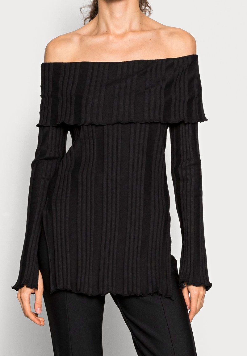 Žena má na sobě černý žebrovaný top s dlouhým rukávem a širokými manžetami, který odhaluje ramena, doplněný o černé kalhoty. Top má mírně volánkový lem.