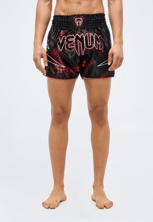 Άνδρας που φοράει μαύρα και κόκκινα σορτς Muay Thai με τη μάρκα Venum και σχέδιο φολιδωτής φίδιου, όρθιος ξυπόλυτος σε λευκό φόντο.