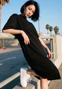 Ung kvinde i sort kjole og hvide sneakers, der læner sig op ad et metalgelænder udendørs med palmer og blå himmel i baggrunden.