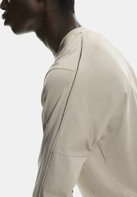 Cămașă bej cu mânecă lungă, ajustată, cu detalii discrete de cusătură pe umeri și mâneci, realizată dintr-un material fin.