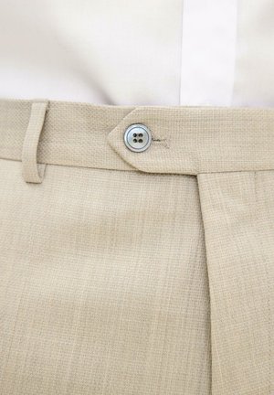 Beige, gestructureerde jurkbroek met een zilveren knoop met vier gaten, dichtgemaakt bij de taille, gedragen met een wit, ingestopt overhemd.