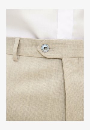 Beżowe spodnie z teksturowanego materiału zapięte na srebrny guzik z czterema dziurkami w talii, noszone z białą włożoną koszulą.