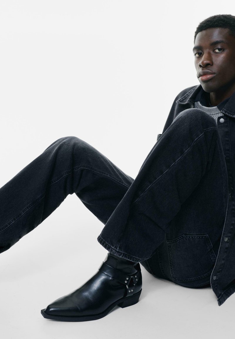 Jachetă din denim negru și pantaloni cu o croială lejeră, asortați cu cizme negre din piele până la glezna, punctate cu accente decorative din metal.