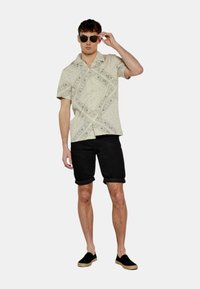 Camicia a maniche corte di colore beige chiaro con motivo e dettagli scuri, abbinata a pantaloni corti neri arrotolati e scarpe slip-on nere, modello con occhiali da sole.