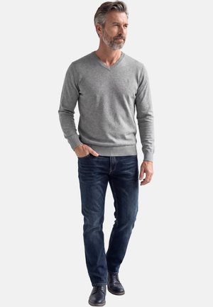 Mann trägt grauen V-Ausschnitt-Pullover, dunkelblaue Jeans und schwarze Lederschuhe, steht mit einer Hand in der Tasche und schaut zur Seite.