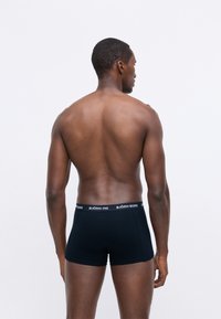 Skaistuma vīrietis stāv ar muguru pret kameru, valkājot melnas Björn Borg bokserbikses uz vienkrāsaina baltā fona.