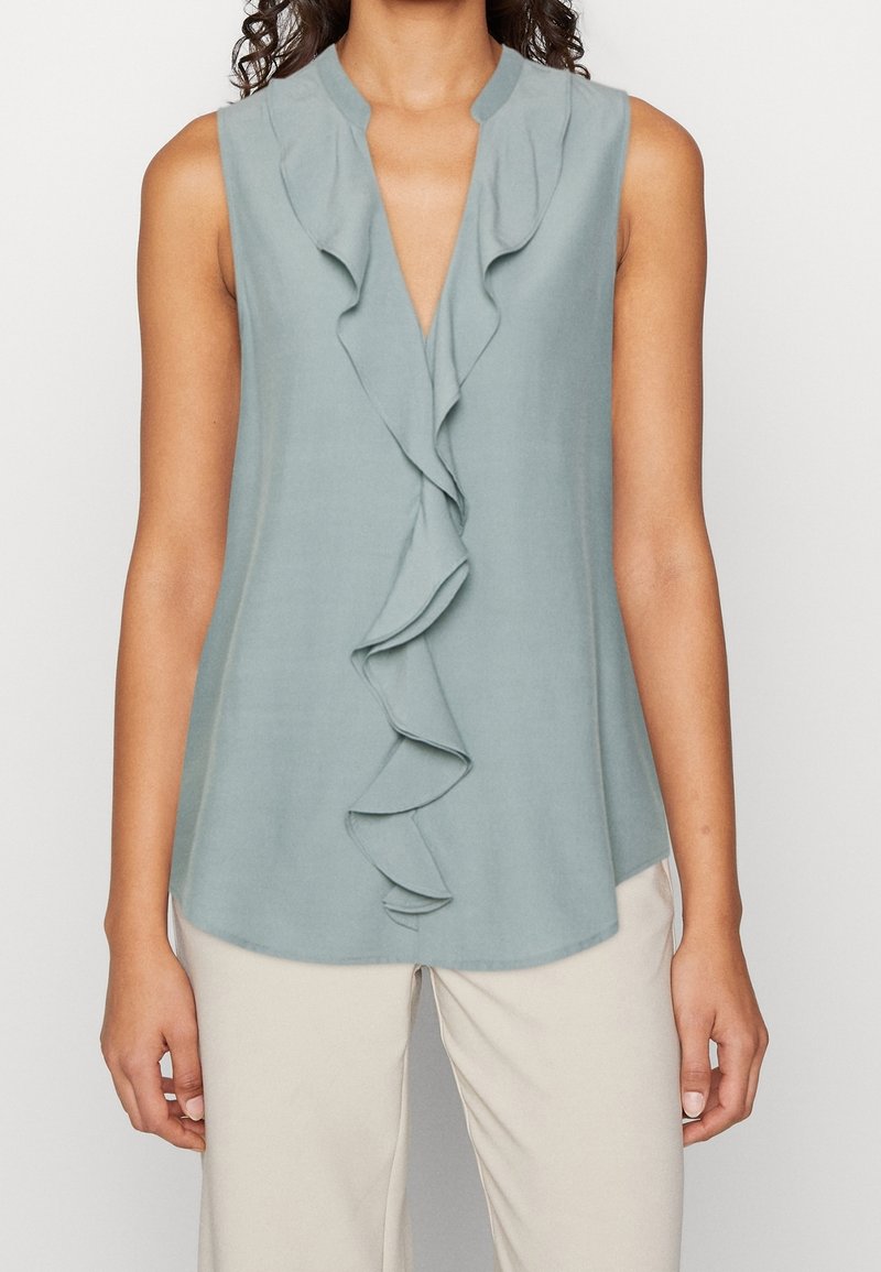 Haut sans manches bleu clair avec un pli froncé sur le devant, un col en V et une texture lisse. Associé à un pantalon beige.