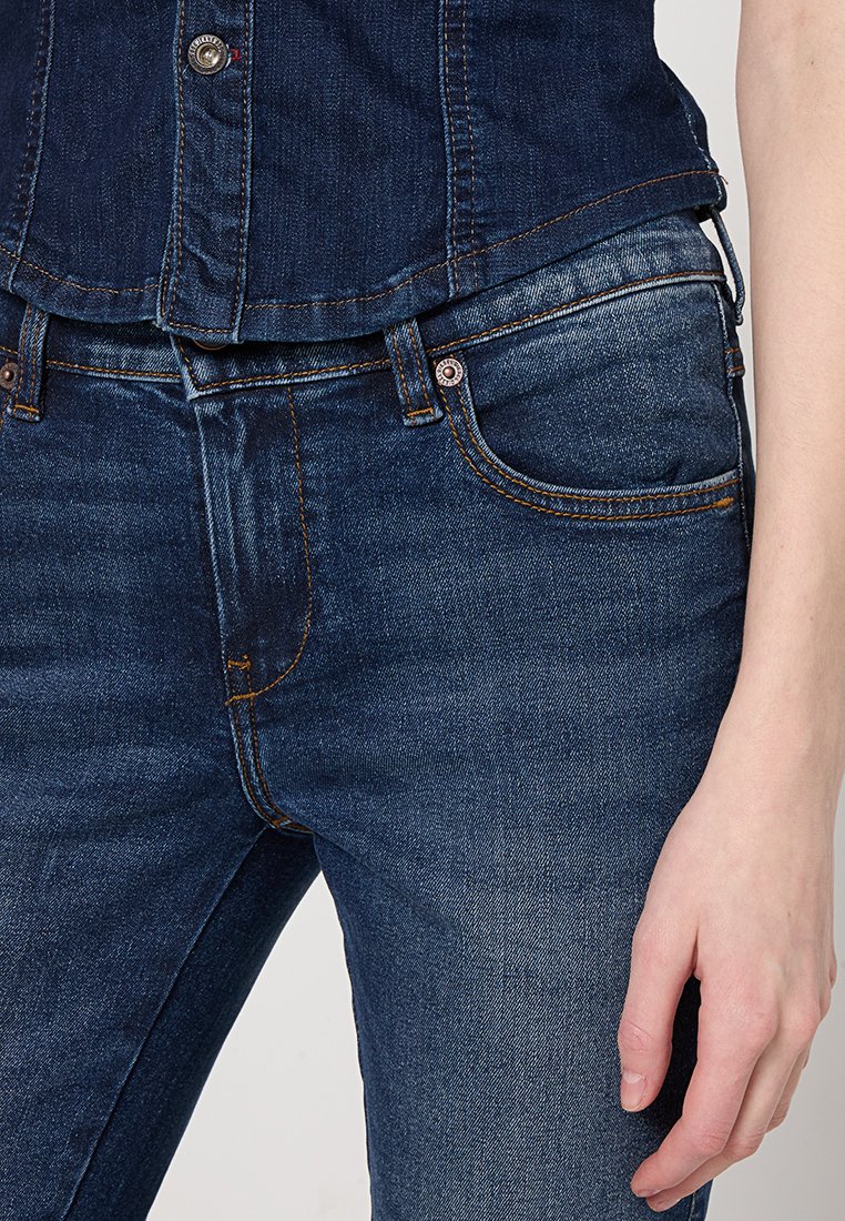 Donkerblauwe spijkerbroek met een getailleerde pasvorm, contrasterende stiksels, vijf zakken en een metalen knoopsluiting zichtbaar bij de taille.