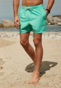 Türkiz színű fürdőnadrág, állítható derékpánttal, oldalsó zsebekkel és logóval. Homokos strandon viselik, háttérben víz látható.