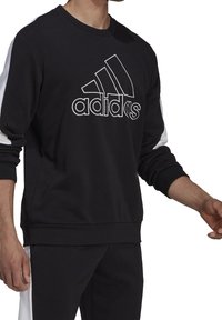 Sweat-shirt noir avec des accents blancs sur les manches, présentant un grand logo Adidas brodé en blanc au centre. Matière en mélange de coton.