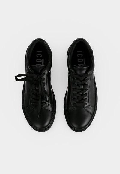 Paire de baskets en cuir noir avec des lacets noirs sur fond blanc, les lacets d'une chaussure sont noués et ceux de l'autre desserrés.