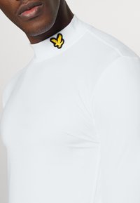 White long-sleeve top with a high collar, featuring a black and yellow eagle emblem on the left shoulder. Smooth fabric texture.