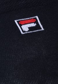 Black fabric with a textured finish featuring a red, white, and black Fila logo in a rectangular shape, embroidered on the surface.
