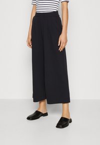 Svarta vida byxor med veck, tillverkade av mjukt material. Kombineras med svarta slip-on skor och en vit randig kortärmad tröja.