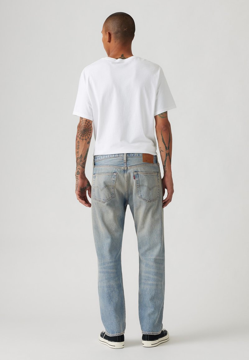 Weißes Kurzarm-T-Shirt kombiniert mit hellblauen Jeans. Die Jeans haben eine ausgewaschene Optik, einen geraden Schnitt und zwei Gesäßtaschen. Schwarze Sneaker sind sichtbar.