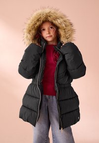 Černá blesková bunda s kapucí lemovanou umělým kožíškem, prošívaným vzorem, elastickým pasem a předním zipem. Nositelná přes vínové tričko a světle modré džíny.