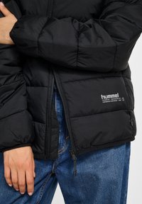 Zwarte pufferjas met doorgestikt ontwerp, voorzien van een ritssluiting aan de voorkant en logodetail. Draagt over blauwe spijkerbroek met een gladde textuur.