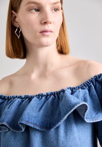 Denim majica bez rukava s detaljima nabranog izreza i elastičnog trimanja. Model nosi srebrnu apstraktnu naušnicu. Meka tekstura, plava boja.