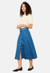 Jeans maxi-rok met een voorknoopsluiting, hoge split en een blauwe kleur. Combinatie met een crèmekleurige gebreide top en zwarte ballerina's.