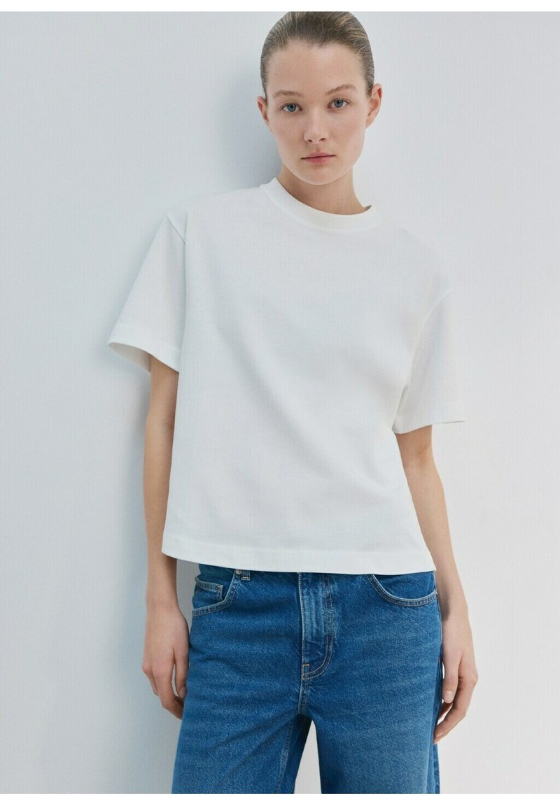 Jeune femme aux cheveux tirés en arrière portant un t-shirt blanc uni à manches courtes et un jean bleu, posant devant un mur blanc.