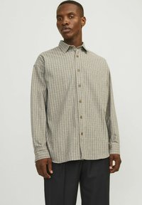 Chemise à carreaux beige avec un motif en grille bleu marine et rouge, à manches longues et boutons marron. Portée par-dessus un pantalon sombre.