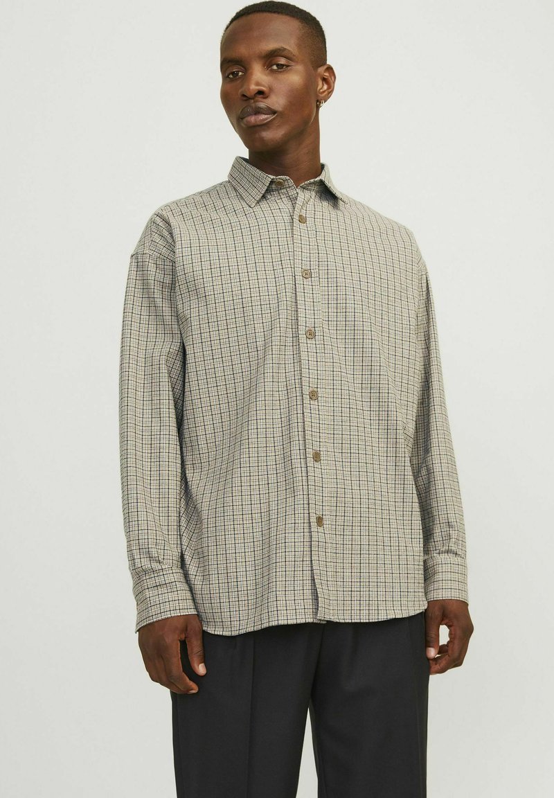 Chemise à carreaux beige avec un motif en grille bleu marine et rouge, à manches longues et boutons marron. Portée par-dessus un pantalon sombre.