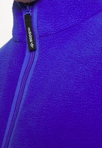 Chaqueta de forro polar morada con cuello alto, cremallera frontal y logo negro de Adidas en el tirador de la cremallera. Textura suave con un tono vibrante.