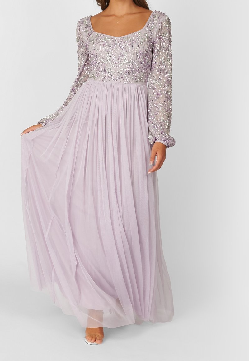 Femme en robe de soirée violet clair à manches longues avec corsage orné et jupe plissée fluide, tenant un côté de la jupe.