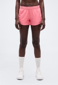 Άτομο που φοράει ροζ αθλητικά σορτς Under Armour, λευκές κάλτσες μέχρι τον αστράγαλο και λευκά αθλητικά παπούτσια, στέκεται μπροστά σε απλό φωτεινό φόντο.