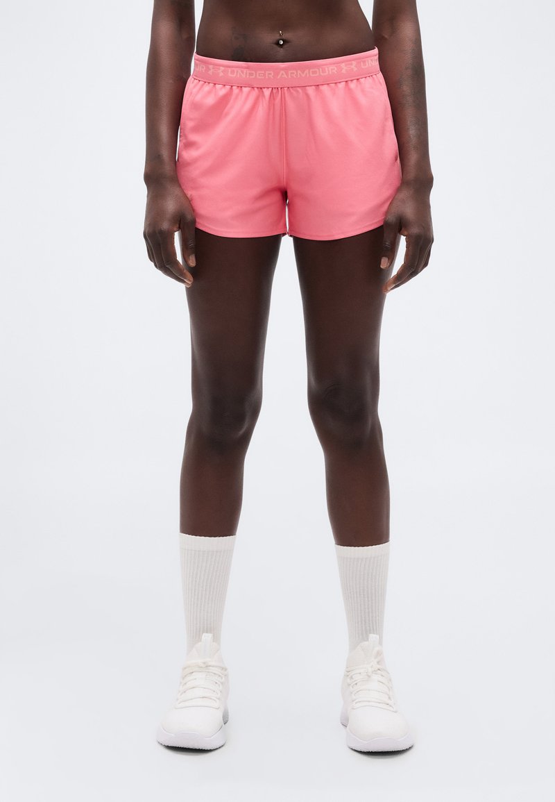 Άτομο που φοράει ροζ αθλητικά σορτς Under Armour, λευκές κάλτσες μέχρι τον αστράγαλο και λευκά αθλητικά παπούτσια, στέκεται μπροστά σε απλό φωτεινό φόντο.