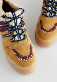 Gelbe Wildleder-Sneaker mit einem strukturierten Fellkragen, navy- und weißgestreiften Schnürsenkeln, roten Akzenten und dekorativer Stickdetails.