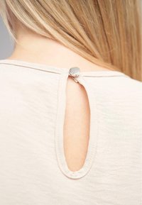 Top beige con un design a apertura chiave sul retro, fissato da un bottone metallico. Tessuto morbido con una finitura leggermente stropicciata.