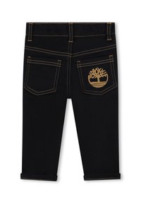 Jeans in denim neri con cuciture dorate a contrasto, design a cinque tasche e logo di un albero dorato sulla tasca posteriore destra. Cuffie arrotolate.