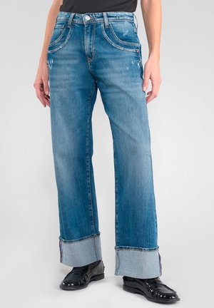 Person trägt blaue, umgeschlagene Jeans mit geradem Bein und schwarze, glänzende Loafers, steht vor einem hellgrauen Hintergrund.