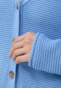Helle blaue Strickjacke mit strukturiertem Muster, Knopfverschluss und gerippten Bündchen. Zeigt eine Hand, die auf der Knopfleiste ruht.