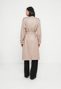 Μπεζ δερμάτινο παλτό  τρεντς με ζώνη στη μέση, επωμίδες στους ώμους και ραγάδες στα μανίκια, σε στυλ με μαύρο παντελόνι και ψηλοτάκουνα.